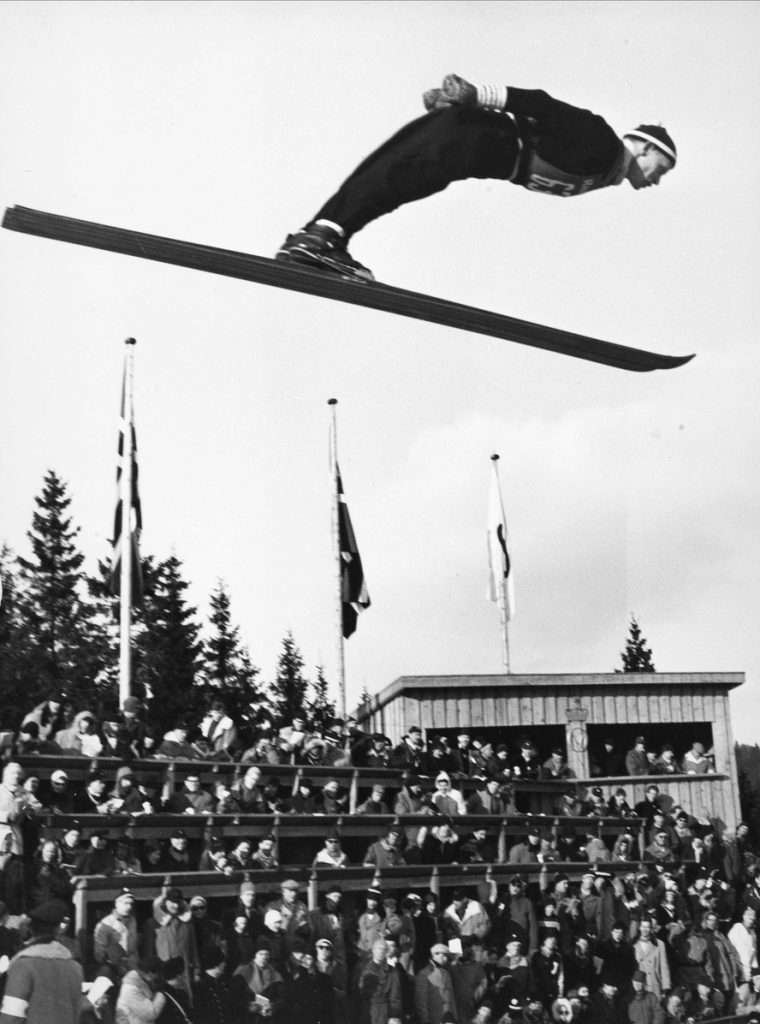 Nordic combined athlete Gunder Gundersen ski jumps with stands of spectators in the background